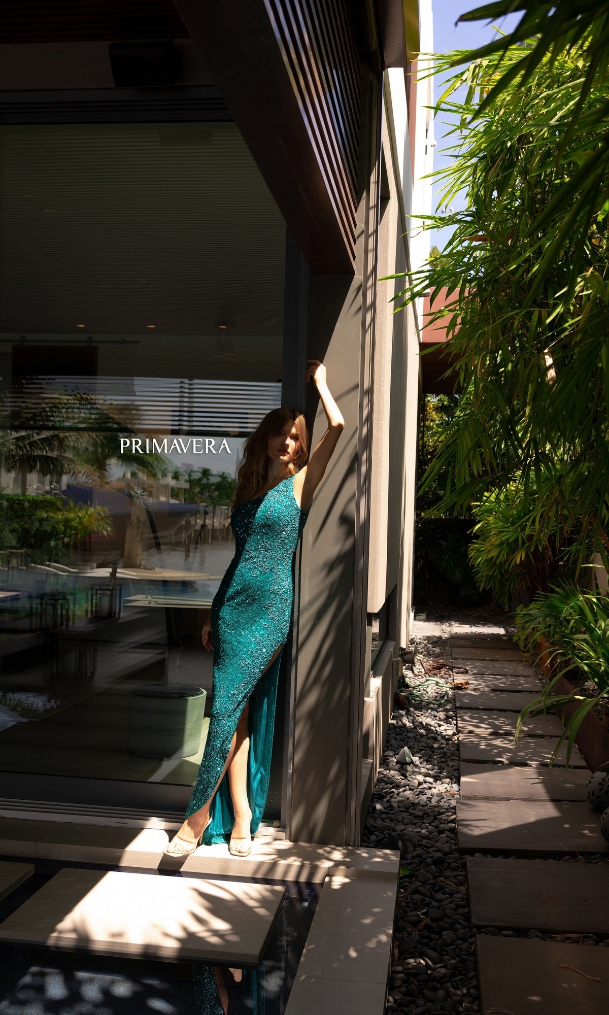 A woman in the Primavera One-Shoulder Long Beaded Prom Dress 4139 poses by a modern building with